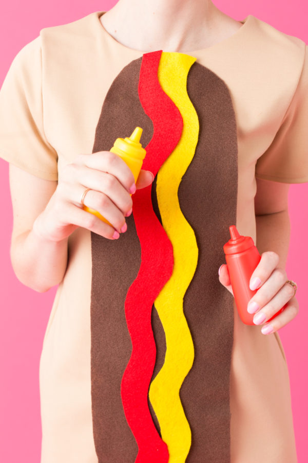 A woman dressed as a hot dog and holding ketchup and mustard.