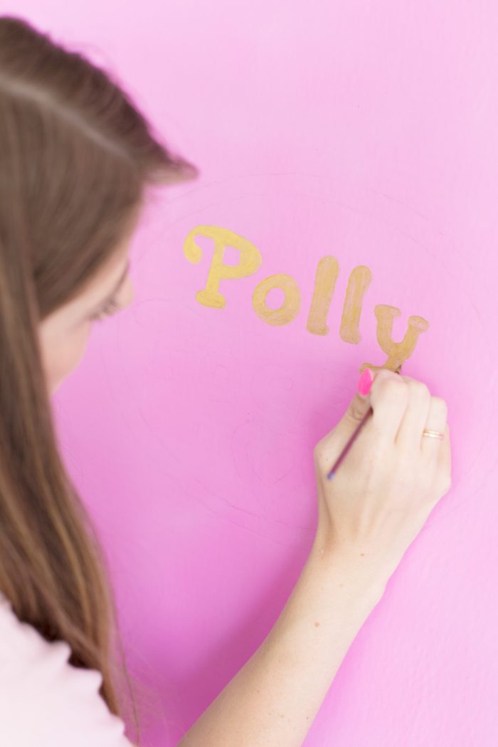 A woman painting the word "polly" in gold on a pink wall