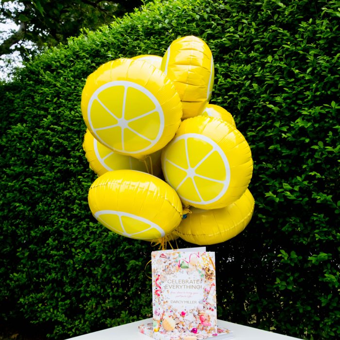 Lemon balloons tied to a table with a green bush in the background. 