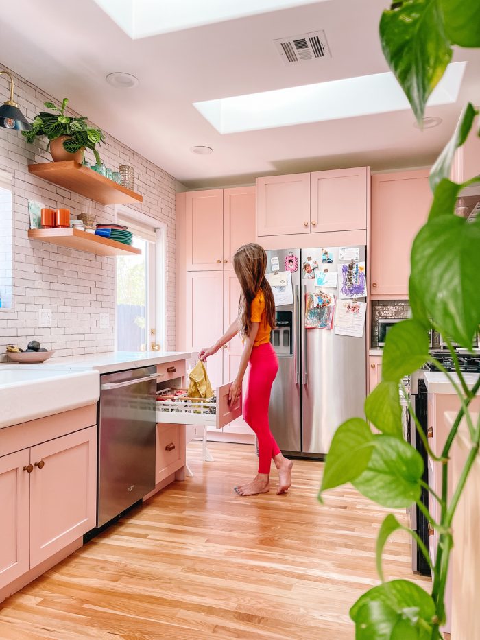 Woman in pink kitchen pulling towel out of drawer with a plant in the foreground