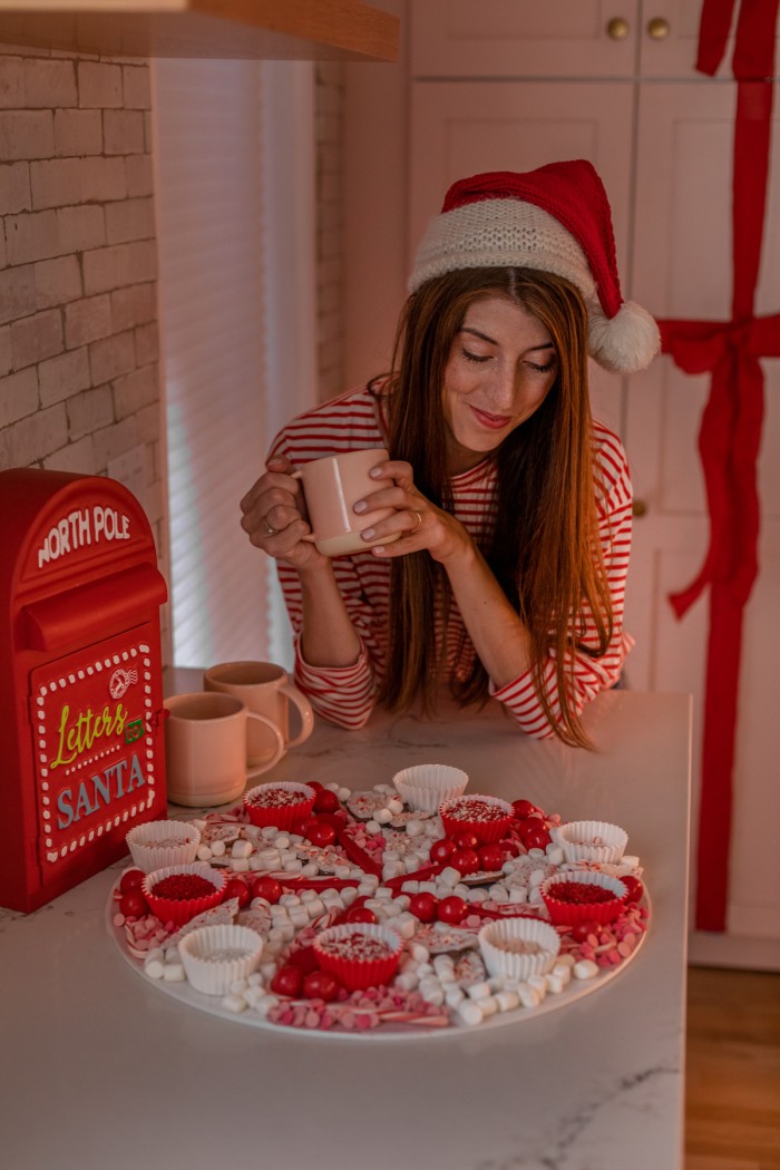 Christmas hot chocolate board with a woman in the background holding a mug and wearing a Santa hat. 