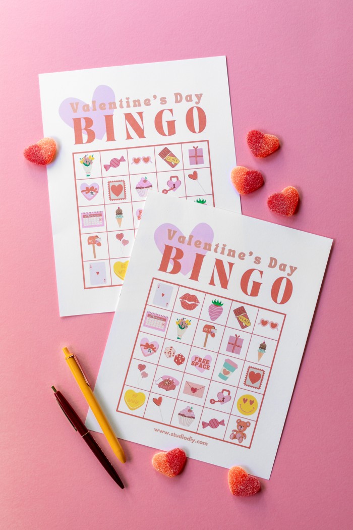 Two Valentine's Day bingo cards next to pens and gummy candies on a pink background. 