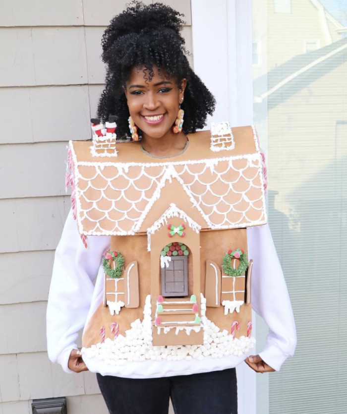 Woman standing in front of a house while wearing a cardboard gingerbread house. 