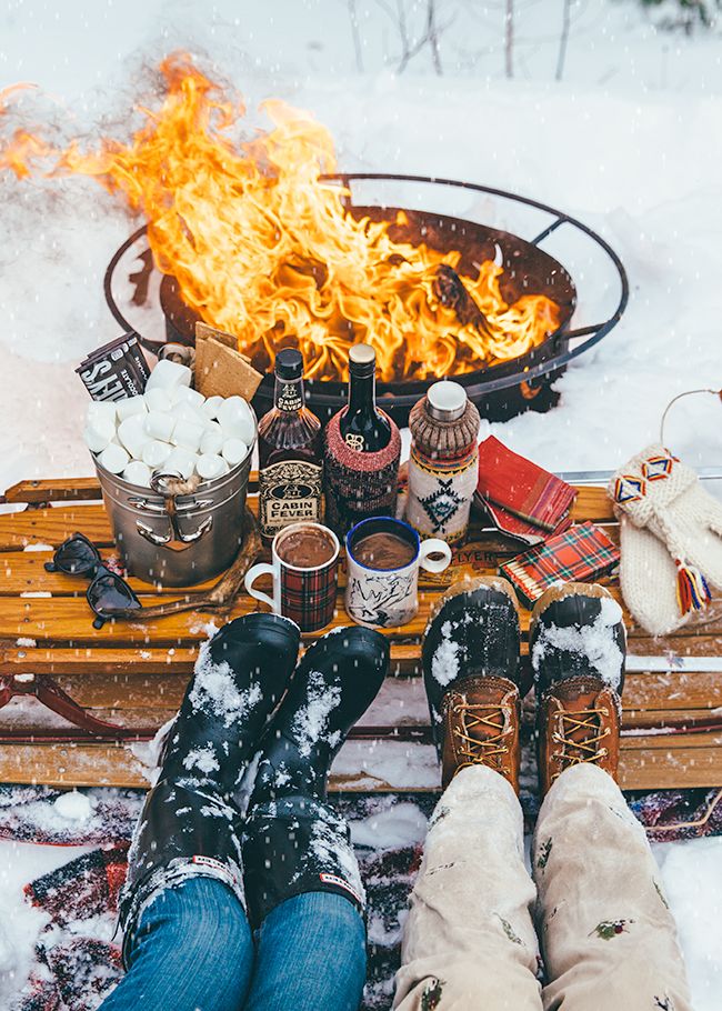 Two pairs of feet close to a bonfire with a bench that has marshmallows and hot chocolate on it. 