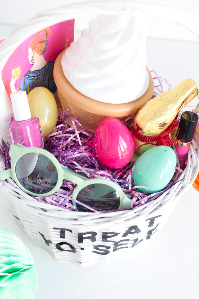 White basket filled with purple shredded paper with sunglasses, nail polish, lipstick and chocolate in the basket. 