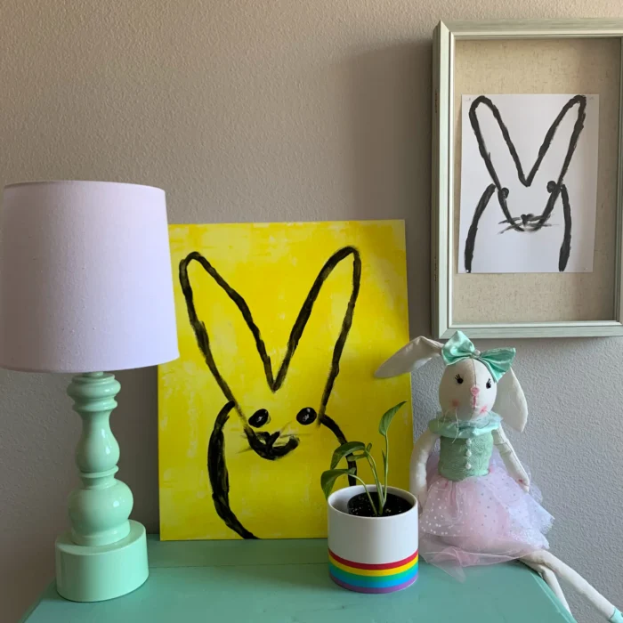 Two paintings of bunnies on a wall next to a night stand. 
