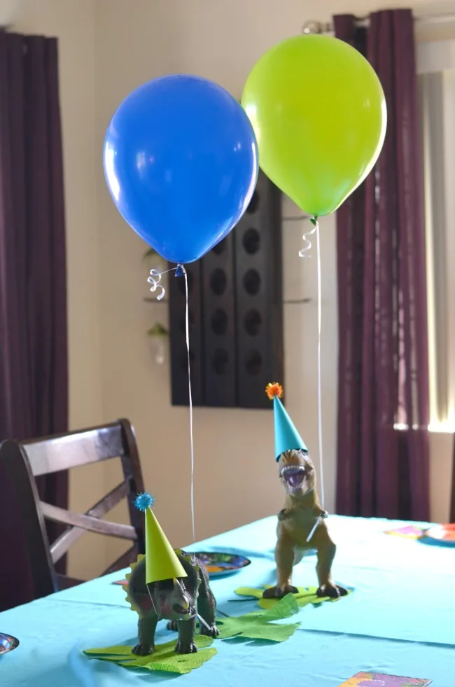 Two dinosaur figures on a table with party hats and balloons. 