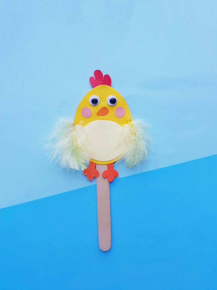 Chick made out of construction paper and glued to a wooden stick on a blue background. 