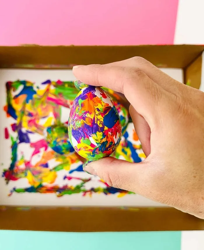 Hand holding an egg covered in paint with a cardboard box full of paint in the background. 