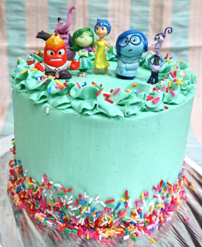 Blue cake with rainbow sprinkles and Inside Out character figures on top. 