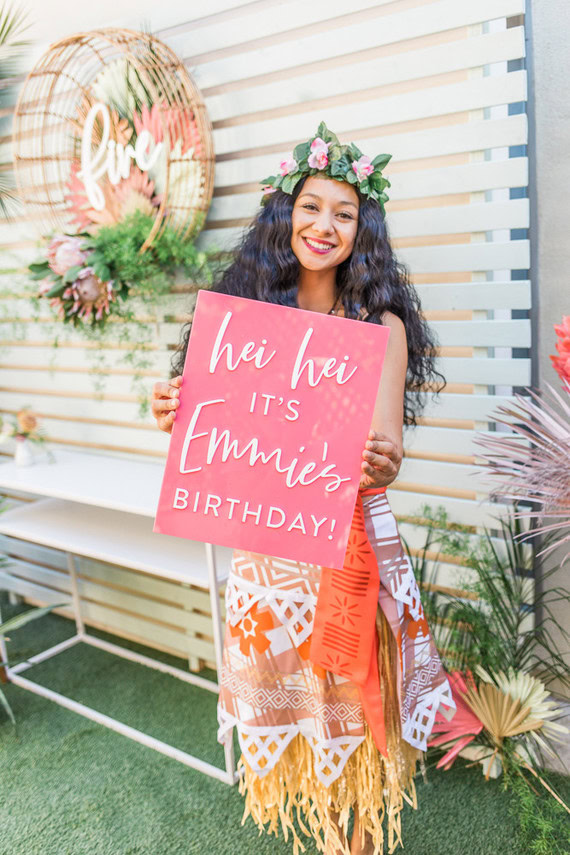 Person dressed up as Moana holding a sign that says "hei hei it's emmie's birthday". 