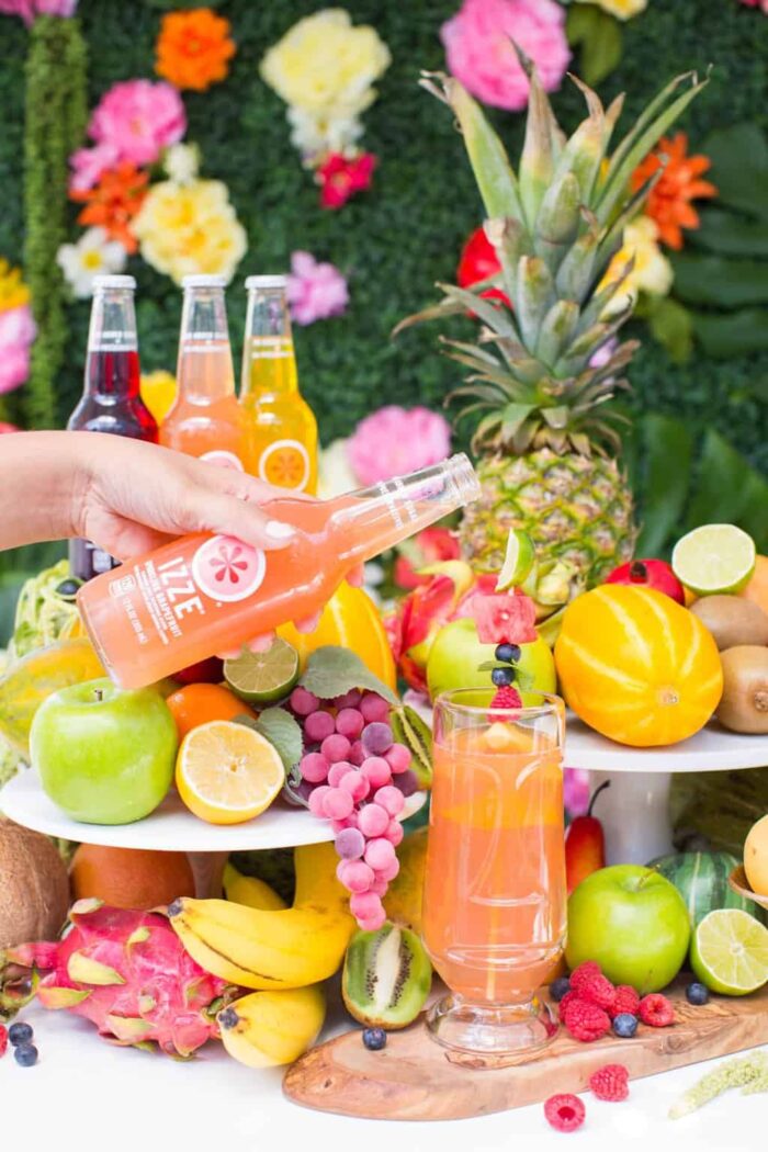Hand holding a bottle of soda and pouring it into a glass surrounded by tropical fruits. 