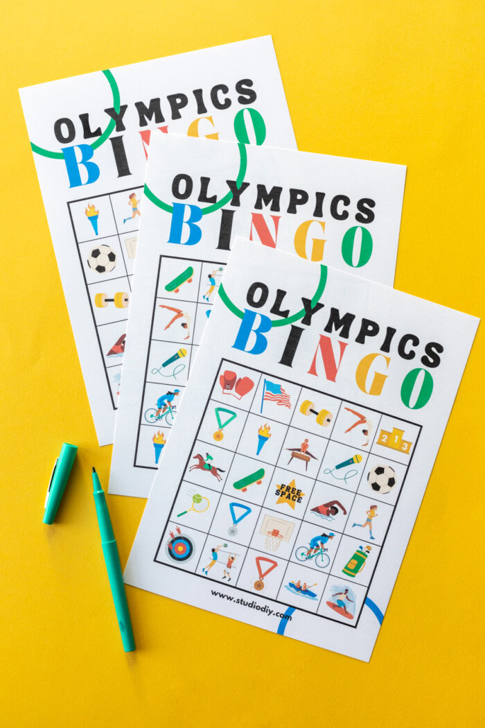 Three Olympics bingo cards on a yellow background next to green markers. 